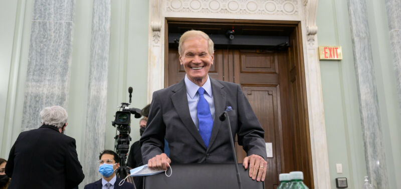 An older man in a suit smiles from a podium.