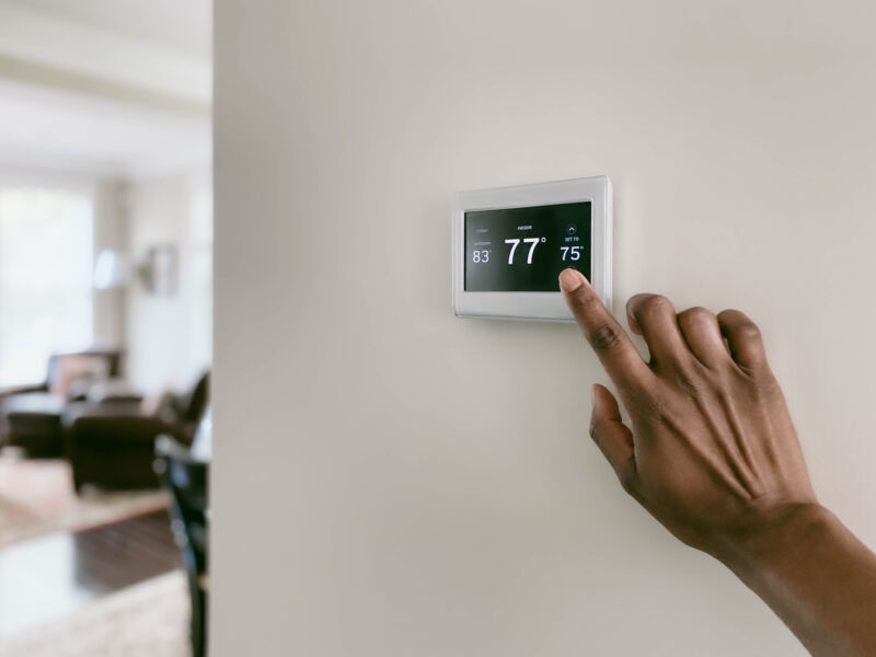 A person's hand adjusting a thermostat that is set to 77° F.