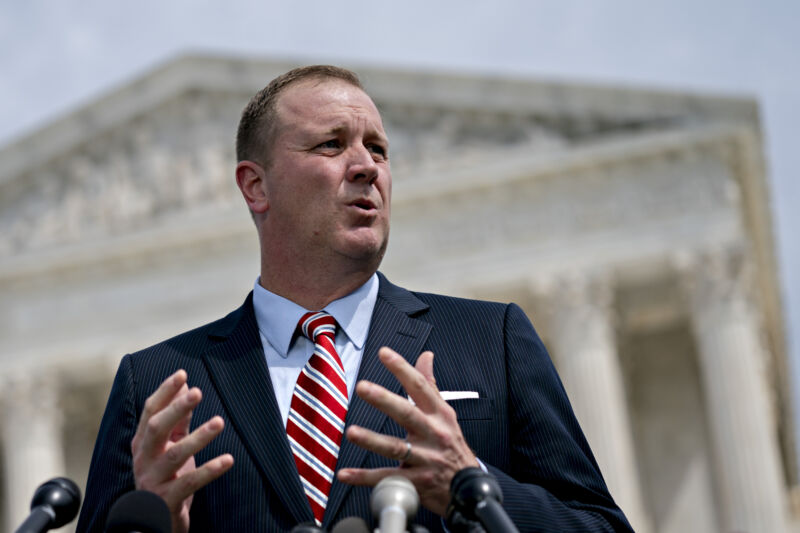 A man in a suit speaks in front of a Neoclassical building.