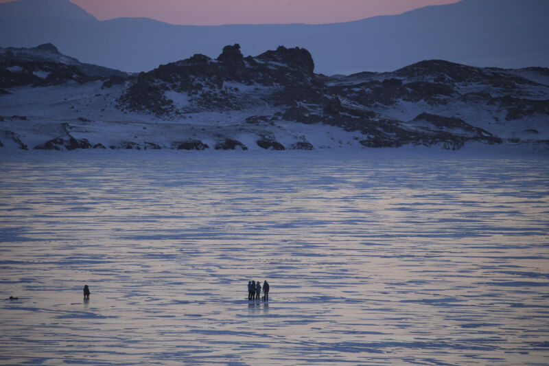 Tiny figures walk on a frozen lake at sunset or sunrise.