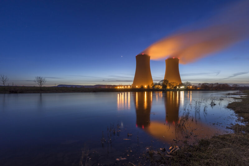 Image Of Two Cooling Towers Above A Body Of Water.