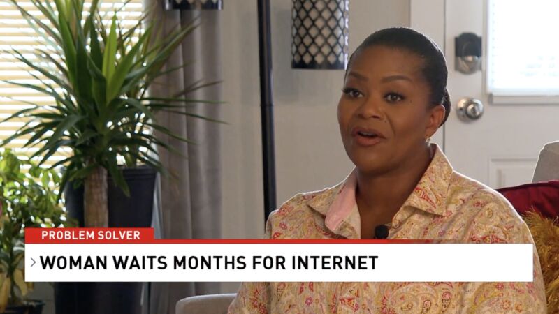 A woman sitting at home and talking to a news reporter. On screen, text says, 