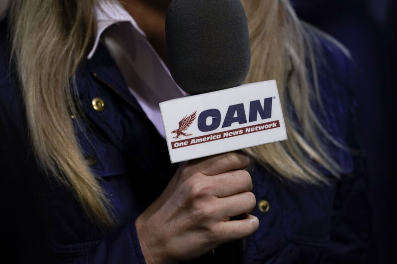 OAN-branded microphone in the hand of a longhaired reporter.