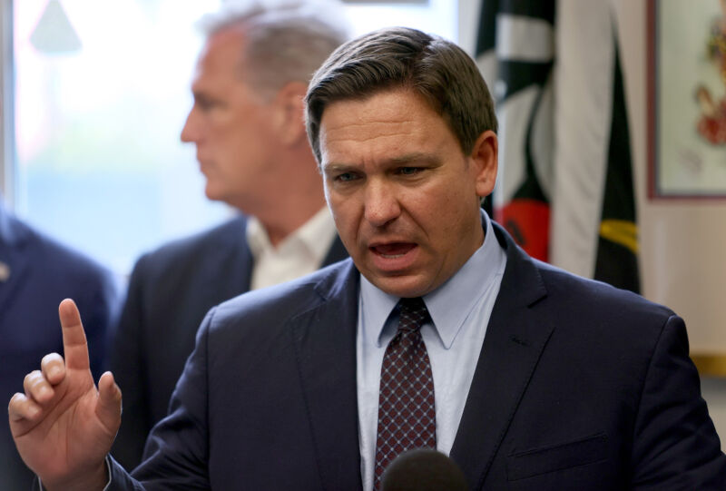 A man in a suit gestures while speaking into a microphone.