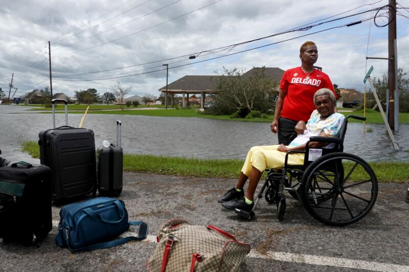 Une femme âgée en fauteuil roulant et sa fille attendent un moyen de transport dans un quartier inondé.