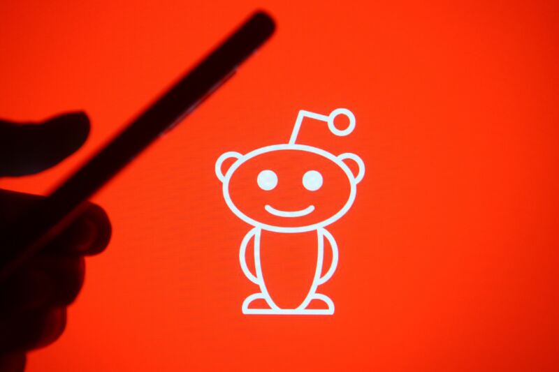 Photo illustration with a hand holding a mobile phone and a Reddit logo in the background.