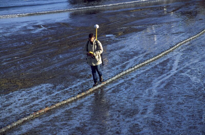 A man walks alongside a cable that runs across a damp, desolate field.