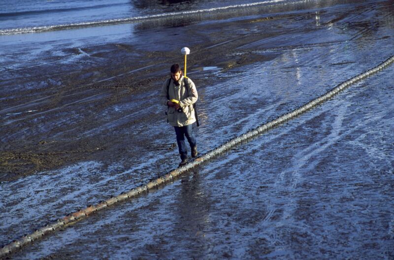 A Man Walks Alongside A Cable That Runs Across A Damp, Desolate Field.