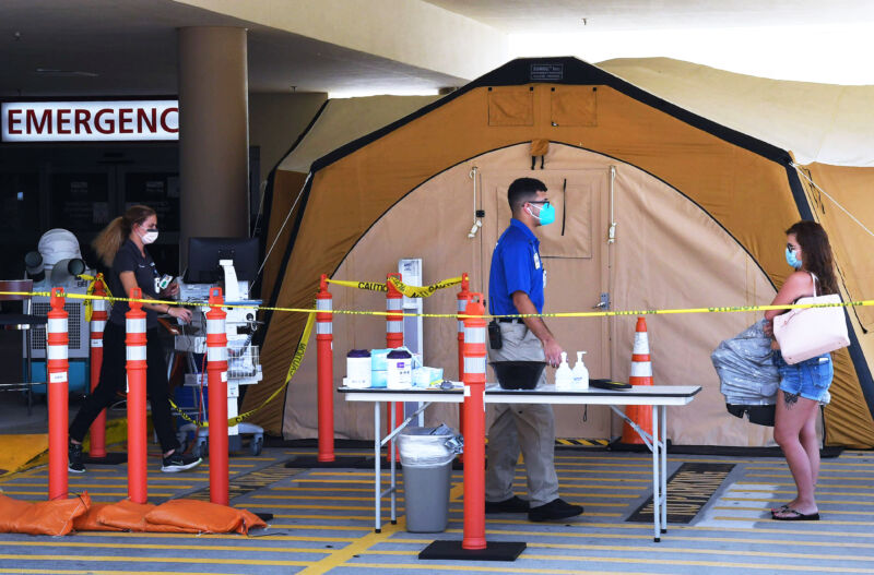 Masked people stand in front of a tent in the middle of traffic cones.