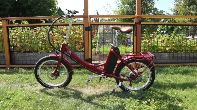 An electric bike with slight unusual proportions on a grassy lawn in front of a fenced garden.