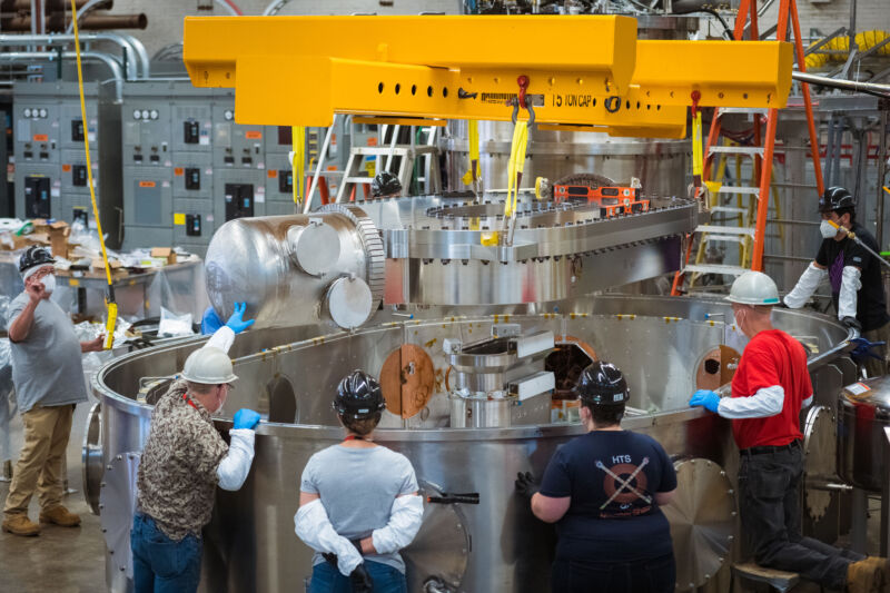 Image of a large metal oval being lowered into a tank by a crane as people observe.