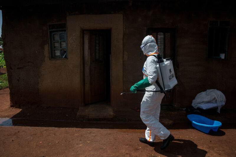 A Woman In Heavy Protective Gear Walks Outside A Small House.