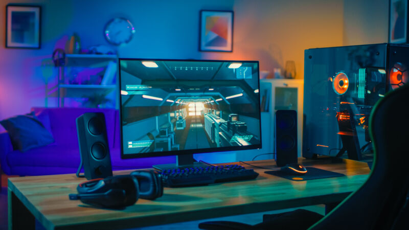 Powerful Personal Computer Gamer Rig with First-Person Shooter Game on Screen. Monitor Stands on the Table at Home. Cozy Room with Modern Design is Lit with Warm and Neon Light.