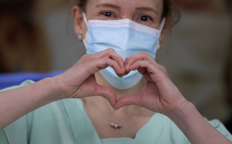 A masked woman makes a heart symbol with her hands.