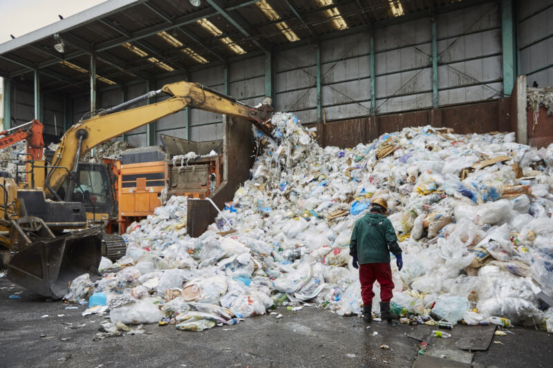 A worker stands in front of a giant mound of plastic waste.