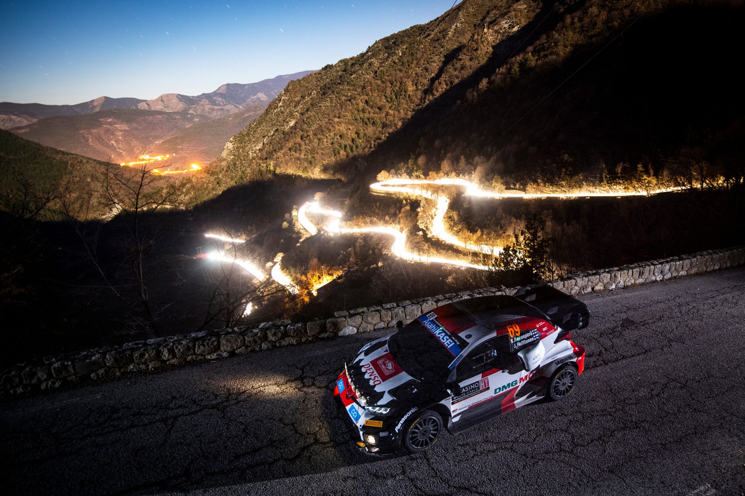 a long exposure composite shot shows a rally car overlooking a road that's glowing gold from time-lapsed headlights.