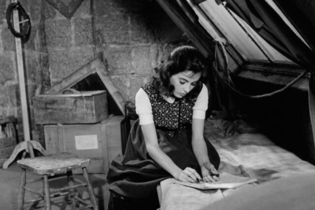 Millie Perkins sits on a bed writing on a tablet of paper in a scene from the 1959 film <em>The Diary Of Anne Frank.</em>