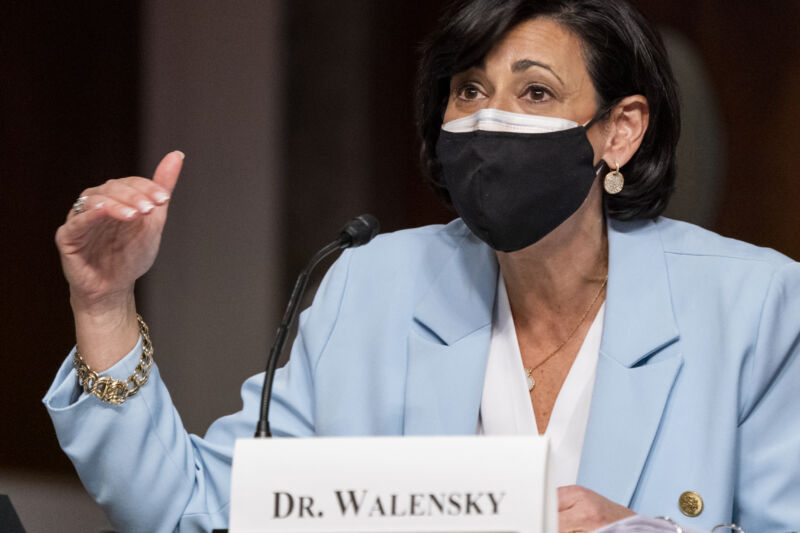 A masked woman in a business suit gestures while speaking into a microphone.
