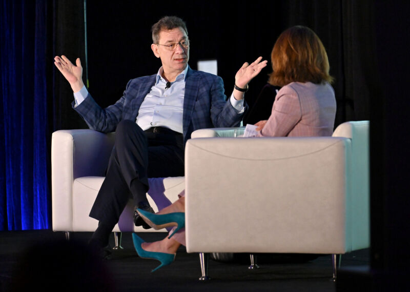 A man in an open-collared suit addresses a woman in a matching chair.