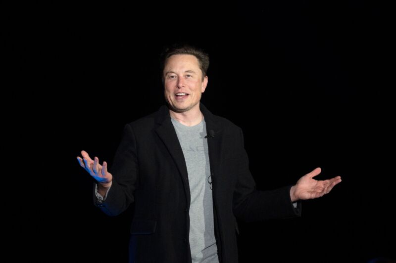 Elon Musk standing and gesturing with his hands while he speaks at a press conference.