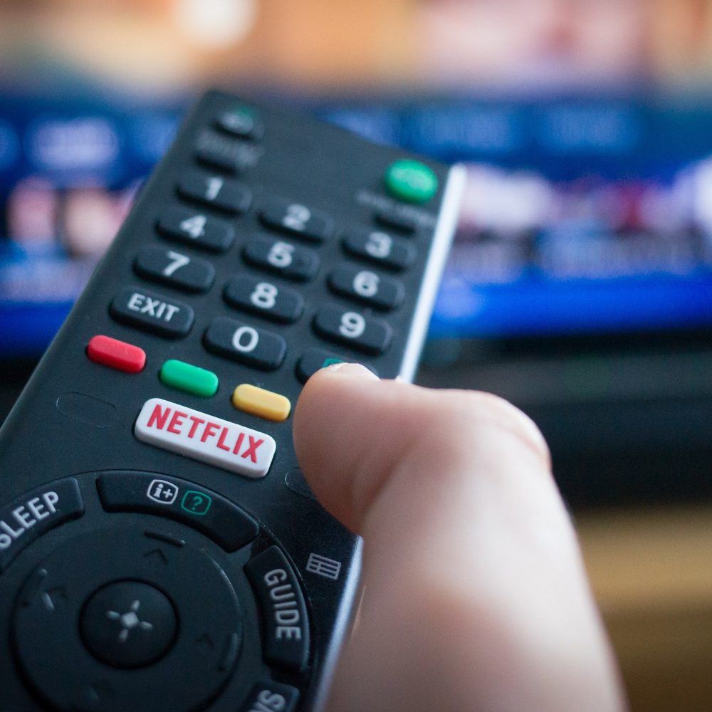 Primer plano de una mano sosteniendo un control remoto de televisión con un botón destacado de Netflix, mientras se observa una pantalla de televisión en segundo plano.