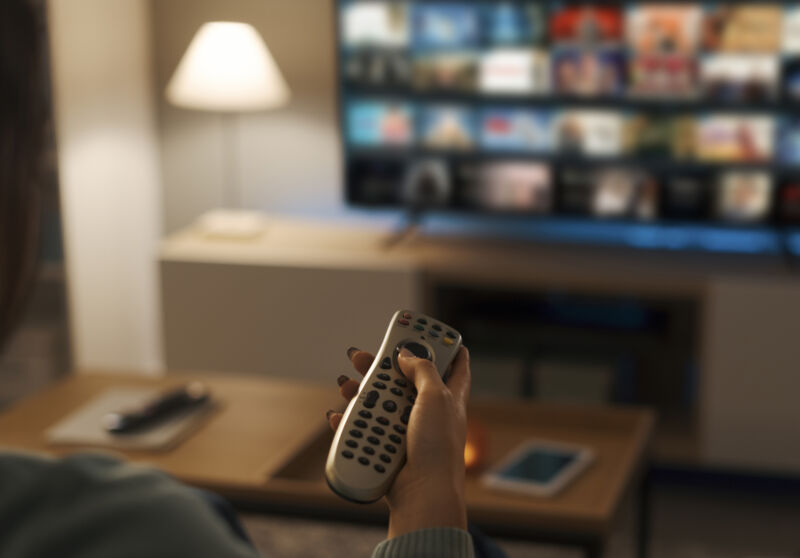 Woman relaxing on the couch and holding a TV remote control while watching videos on demand.