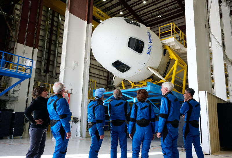 People in blue jumpsuits admire a rocket in a hanger.