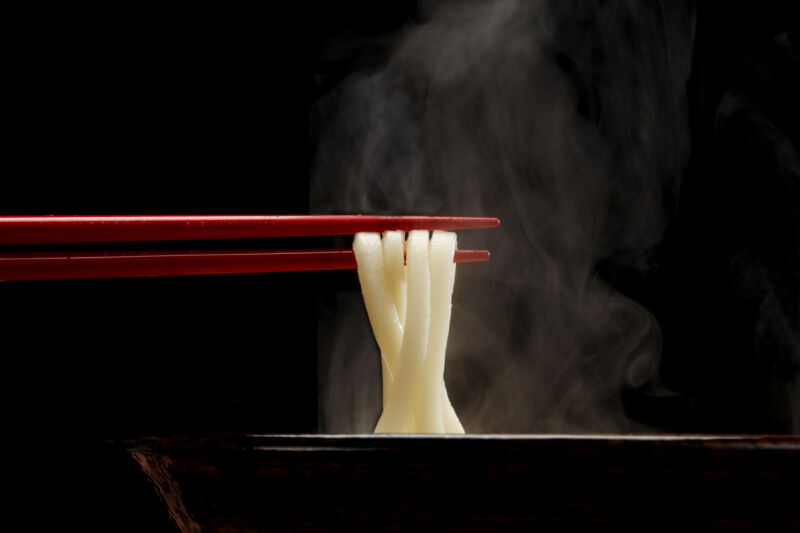 Technology Japanese Sanuki Udon ,fat straight noodle,lifted up with red chopsticks from soup bowl. Steam looks sharp against black background.Eye level angle.