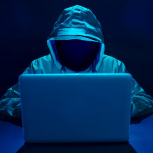 Person wearing a hoodie whose face is not visible sitting in front of a laptop in a dark room.