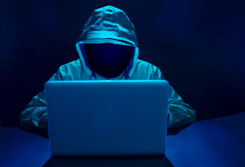 Person wearing a hoodie whose face is not visible sitting in front of a laptop in a dark room.