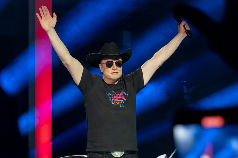 El CEO de Tesla, Elon Musk, en el escenario, sosteniendo un micrófono en una mano mientras ambos brazos están levantados en el aire.