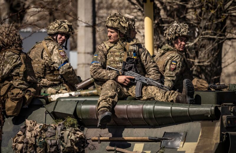 A Ukrainian soldier holding a Kalashnikov-style rifle and other Ukrainian soldiers sit on an armored military vehicle.