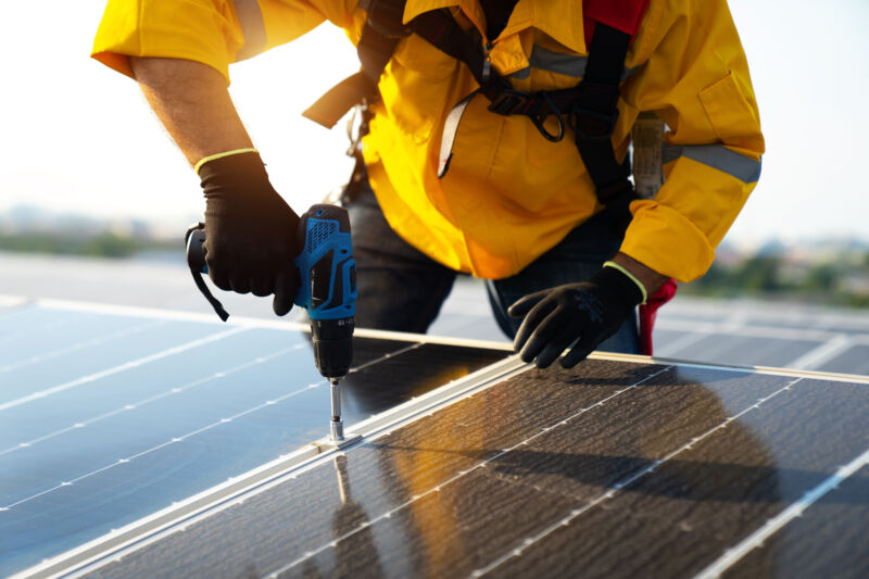 Male technician in yellowsuit installing photovoltaic blue solar modules with screw. Man electrician panel sun sustainable resources renewable energy source alternative innovation