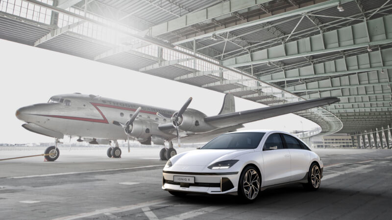 A white Hyundai Ioniq 6 sedan in front of a 1930s airliner in a hangar.