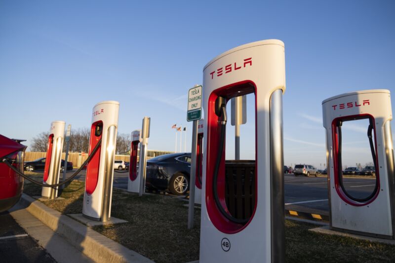 Media docena de cargadores Tesla en un área de estacionamiento, con un par de vehículos Tesla cargándose.