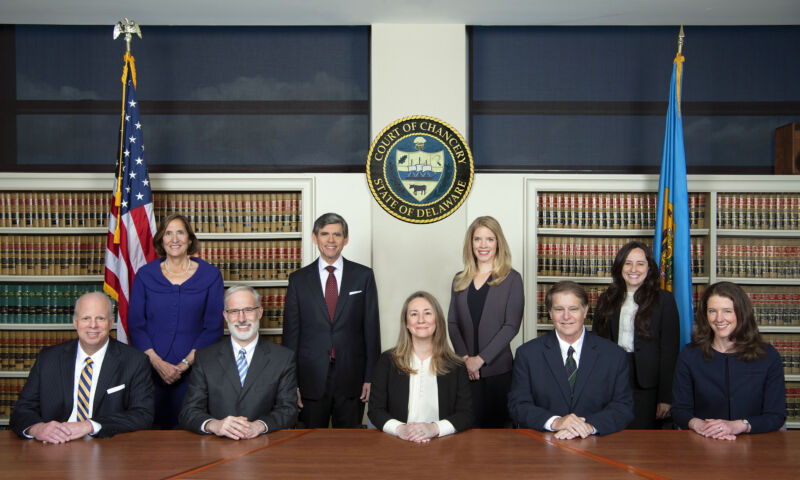 Five judges sit at a table and four judges stand behind them while posing for a picture in front of the Delaware Court of Chancery crest.