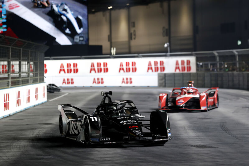 A black formula e car is followed by a red formula e car