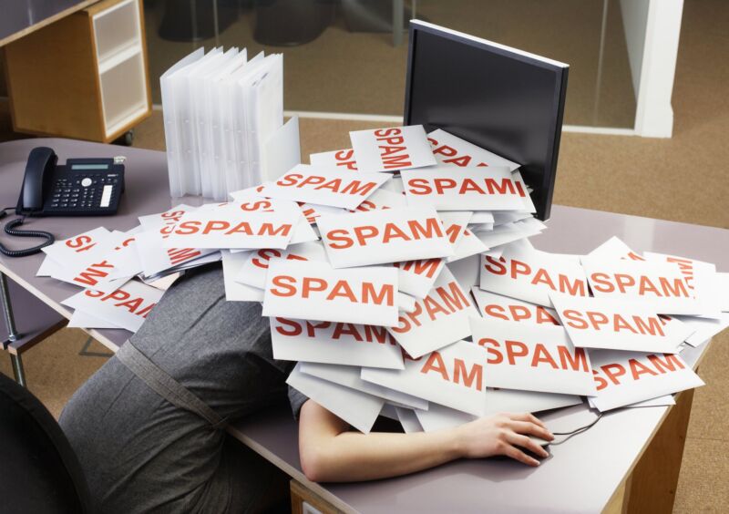 A woman sits at a desk in front of a computer but her head is hidden because she is covered by a massive pile of envelopes labeled