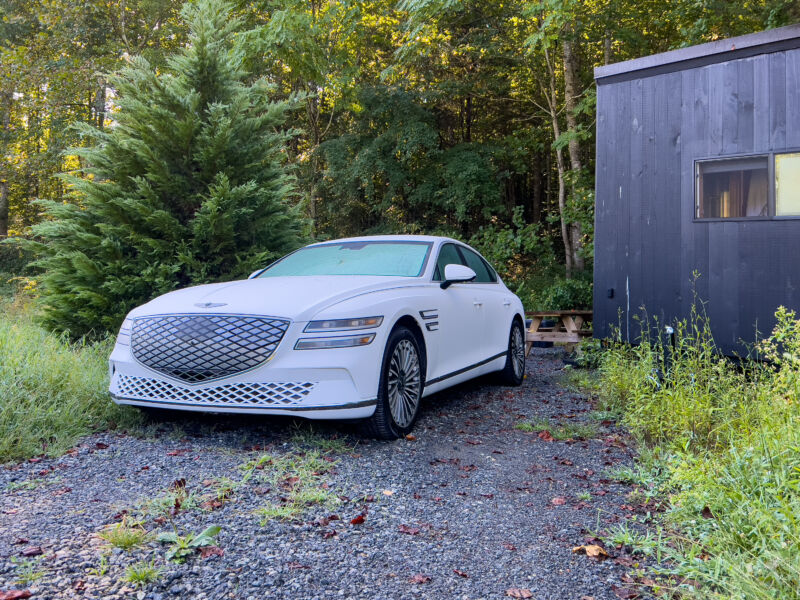 A white Genesis G80 Electrified seen from ahead, next to a small black cabin