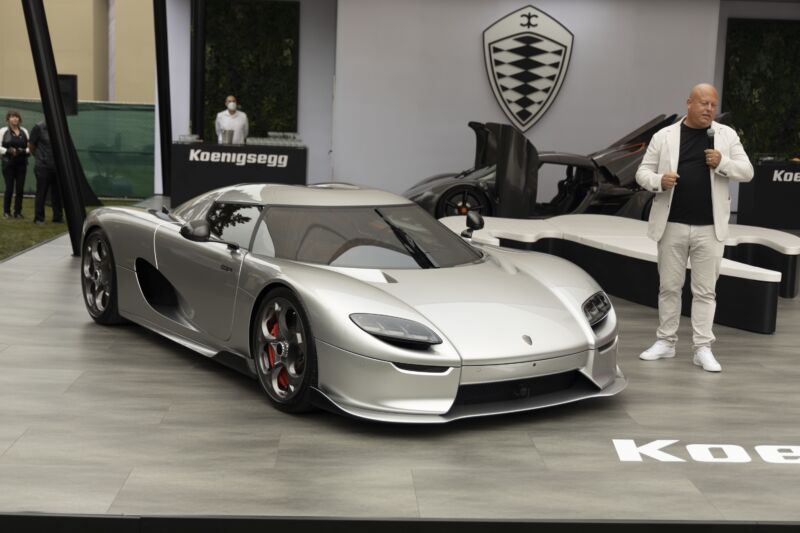 A bald man stands to the right of a silver Koenigsegg supercar