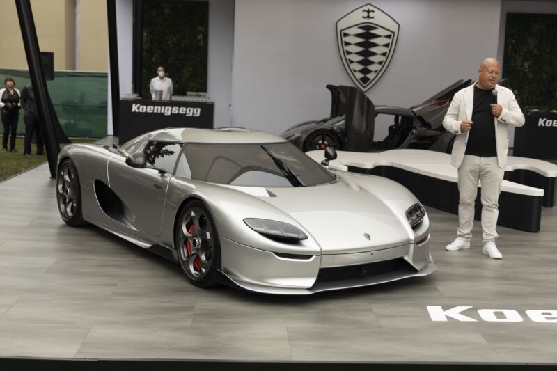 A bald antheral   stands to the close    of a metallic  Koenigsegg supercar