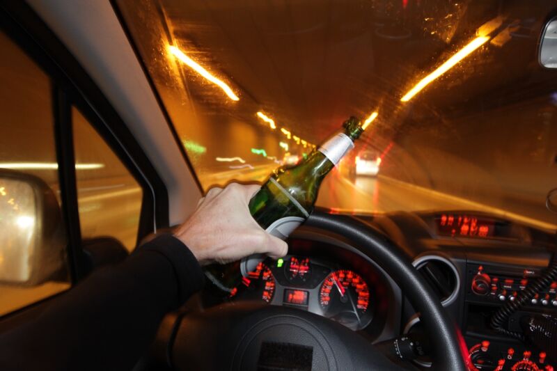 Interior view of a car driving through a tunnel while the driver holds a beer in his left hand, which rests on the steering wheel.