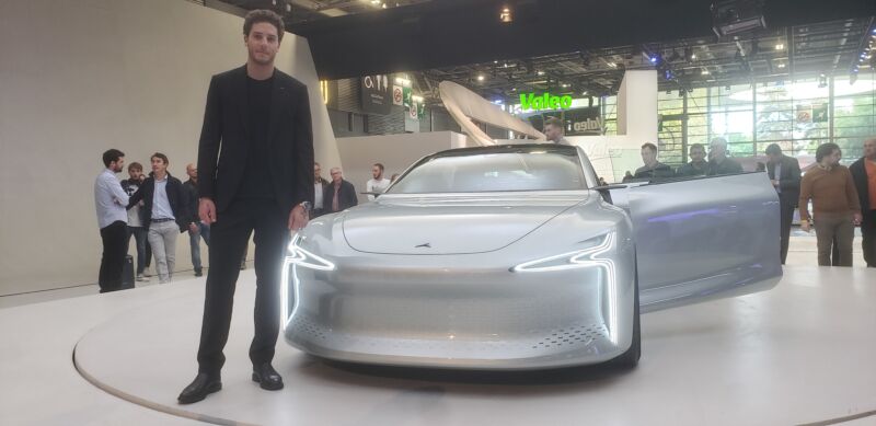 Seorang pria berdiri di samping sedan perak futuristik