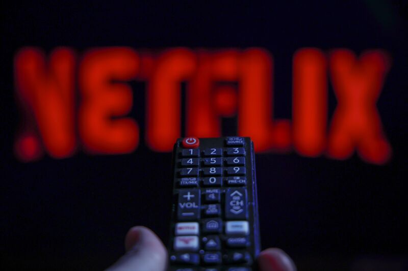 The Netflix logo is shown on a screen while a person holds a TV remote.