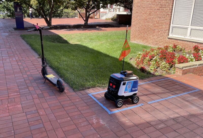 Este robot Kiwibot está esperando que lo carguen con mi muffin.  Luego, un trabajador lo llevó al otro lado de la calle en el scooter.