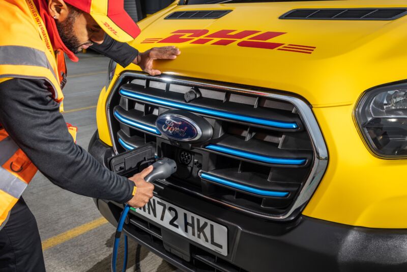 Someone plugs a charger into the front of a Ford E-Transit in DHL colors