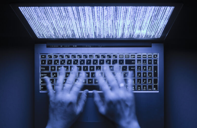 Las manos borrosas están escribiendo en una computadora portátil en la oscuridad con un teclado iluminado y un código de programa místico ilegible visible en la pantalla.