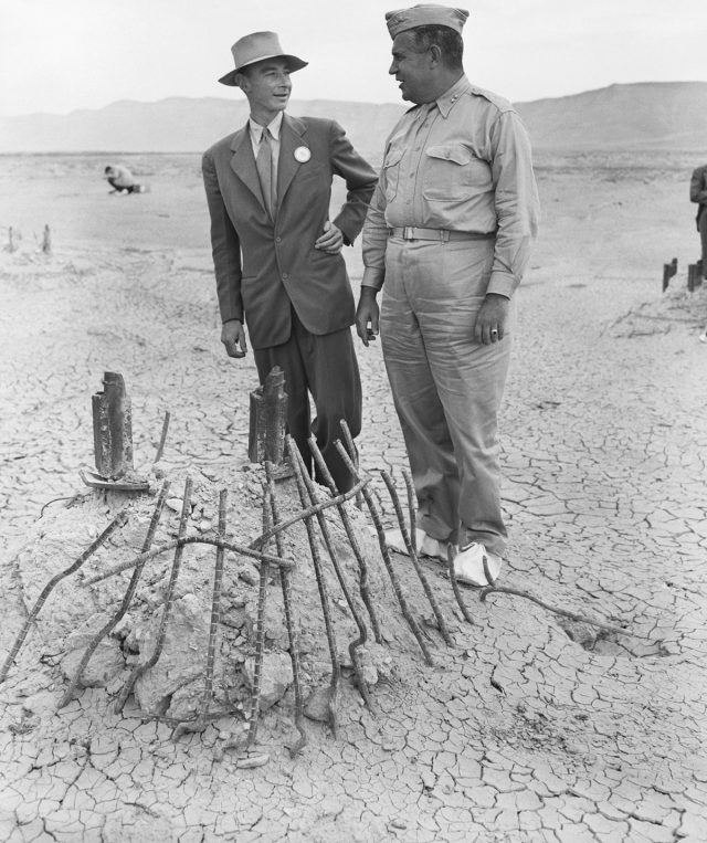 Maj. Gen. Leslie R. Groves (r), and J. Robert Oppenheimer view the base of the steel tower on which the first atomic bomb hung when tested near Alamogordo, New Mexico, in July 1945. The intense heat of the bomb melted the tower, and seared the surrounding sands into jade green glass-like cinders.