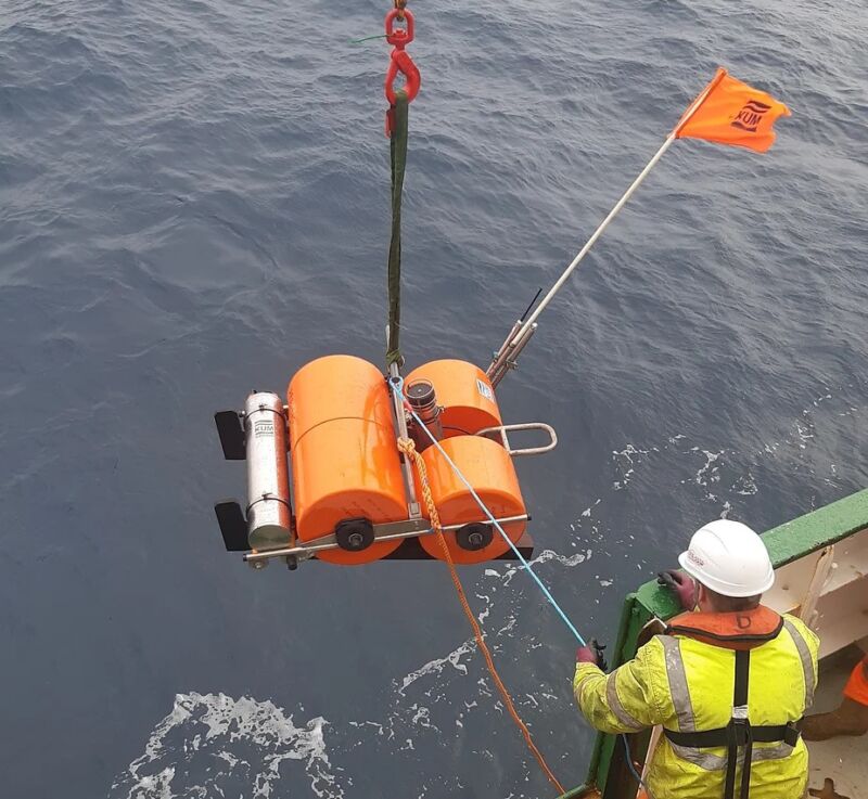 Image of a hard-hatted individual guiding aa large orange device as it's lowered into the ocean.