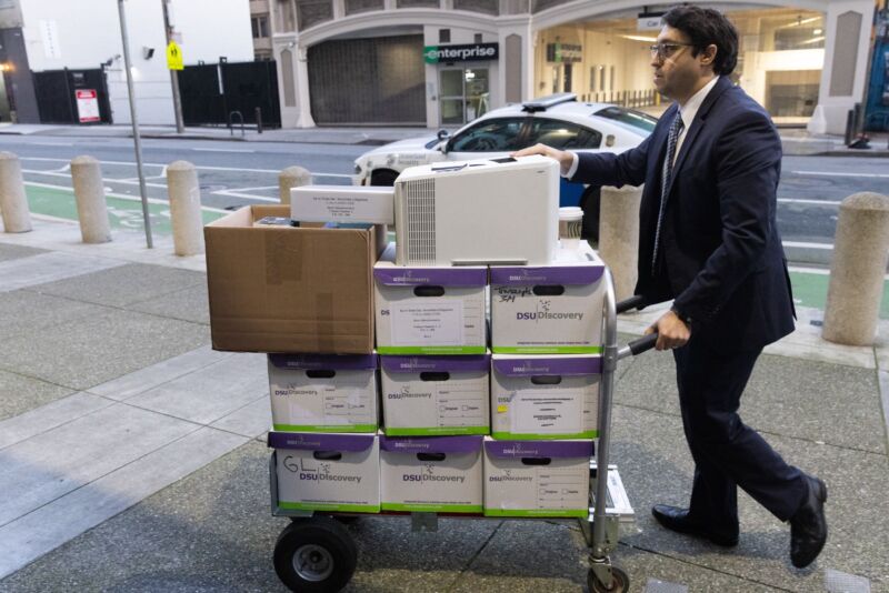 Outside a courthouse, a man pushes a cart holding boxes full of documents related to a trial involving Elon Musk.